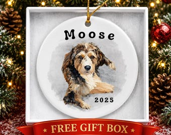 Könnte beinhalten: Ein rundes, weißes Keramikornament mit einer Aquarell-Illustration eines braunen, schwarzen und weißen Hundes. Der Text "Moose" ist schwarz über dem Hund gedruckt, und das Jahr "2025" steht in Schwarz darunter. Ein goldenes Band ist befestigt.