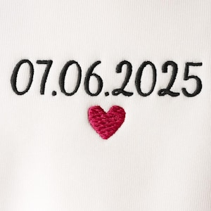 Peut inclure: Tissu blanc avec des chiffres noirs brodés et un cœur rouge. Les chiffres indiquent "07.06.2025". Un cœur rouge est brodé sous les chiffres. Le tissu a une texture subtilement côtelée.