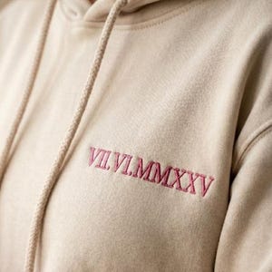 May include: A beige hooded sweatshirt featuring a drawstring hood and the embroidered Roman numeral date VII.VI.MMXXV in dark pink thread on the upper chest area. The garment is made of a soft fabric with a visible texture and long sleeves.