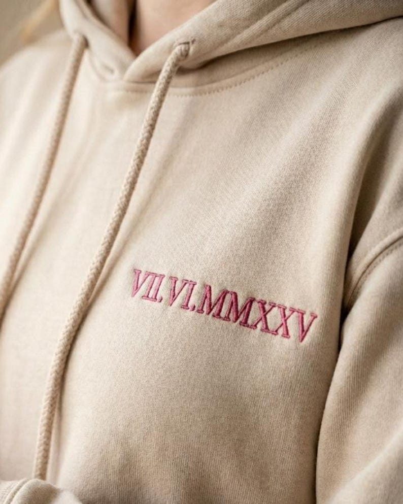May include: A beige hooded sweatshirt featuring a drawstring hood and the embroidered Roman numeral date VII.VI.MMXXV in dark pink thread on the upper chest area. The garment is made of a soft fabric with a visible texture and long sleeves.