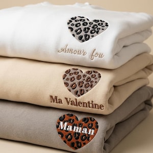 Può includere: Una pila di tre felpe bianche, beige e grigie. Ogni felpa presenta un motivo a cuore leopardato. La felpa bianca ha il testo "Amour fou", quella beige "Ma Valentine" e quella grigia "Maman".
