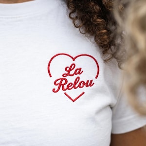 Peut inclure: Un t-shirt blanc avec un contour de cœur rouge brodé sur la poitrine. À l'intérieur du cœur, le texte La Relou est cousu dans une police cursive rouge. Le motif est centré sur la partie supérieure avant du vêtement, avec des lignes nettes.