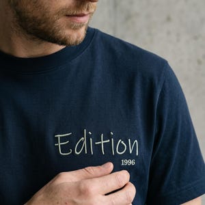 Peut inclure: Un t-shirt bleu marine à manches courtes avec le mot Edition brodé en fil vert sauge clair sur la poitrine, accompagné de l'année 1996 brodée dans une police plus petite et assortie en vert sauge clair juste en dessous du mot.