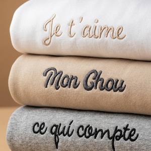 Peut inclure: Trois sweat-shirts pliés, empilés les uns sur les autres, de couleurs blanche, beige et grise. Chaque sweat-shirt porte une broderie en français : "Je t'aime", "Mon Chou" et "ce qui compte". La broderie est assortie à la couleur du sweat.