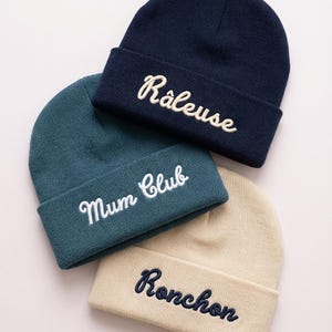 Op de afbeelding: Drie gebreide mutsen in marineblauw, teal en beige. Elke muts heeft geborduurde tekst: "Râleuse", "Mum Club" en "Ronchon". De mutsen zijn omgeslagen aan de rand en zijn gemaakt van zacht, gebreid materiaal.