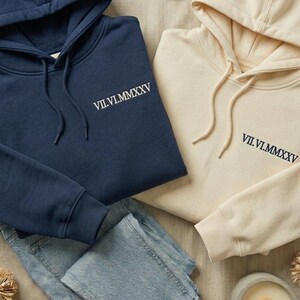 May include: A navy blue hoodie and a cream-colored hoodie, both featuring the Roman numeral date VII.VI.MMXXV embroidered on the chest. Each hooded sweatshirt includes matching drawstrings, long sleeves with ribbed cuffs, and a front kangaroo pocket.