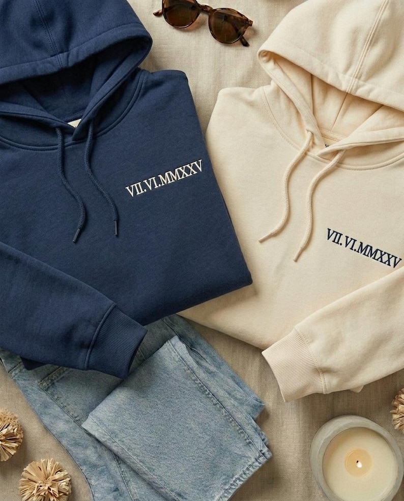 May include: A navy blue hoodie and a cream-colored hoodie, both featuring the Roman numeral date VII.VI.MMXXV embroidered on the chest. Each hooded sweatshirt includes matching drawstrings, long sleeves with ribbed cuffs, and a front kangaroo pocket.