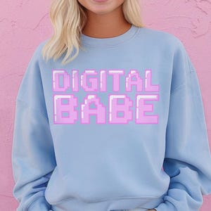 May include: A pale blue crew neck sweatshirt featuring the pink text "DIGITAL BABE" in a pixelated font. The sweatshirt is worn with denim shorts. The background is a solid pink colour.