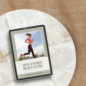 May include: A digital tablet displays a woman running in athletic wear under a blue sky. The tablet's screen features the text "MINI ENERGY RESET GUIDE" and "RESTORE MY SOUL HEALTHCARE". The tablet rests on a marble-like surface, with a burlap background.