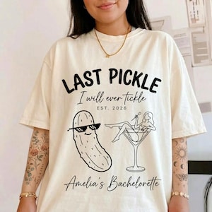 May include: Cream-colored t-shirt with the text "LAST PICKLE I will ever tickle EST. 2026" in black. Features a cartoon pickle wearing sunglasses and a woman in a martini glass. "Amelia's Bachelorette" is printed at the bottom.