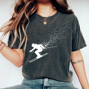 May include: Dark gray t-shirt featuring a white silhouette of a skier in motion, with a trail of white dots trailing behind. The shirt has a crew neck and short sleeves. The design is centered on the front of the shirt.