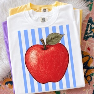 Peut inclure: Un t-shirt blanc à manches courtes avec un motif de grosse pomme rouge dotée d'une feuille verte et d'une tige brune, centré sur un fond de rayures verticales bleu clair. Le t-shirt porte une étiquette Comfort Colors à l'encolure.