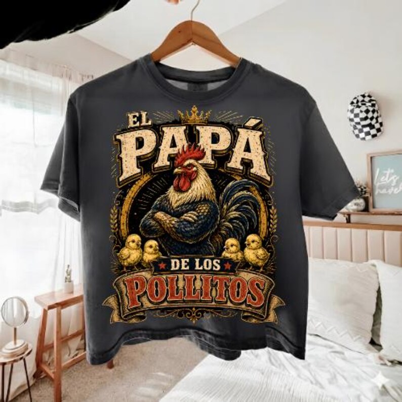 May include: A charcoal gray t-shirt featuring a graphic of a rooster with folded wings wearing a crown, surrounded by four small chicks. The text reads EL PAPA DE LOS POLLITOS in bold, distressed lettering, with a circular border and star accents.
