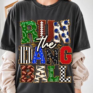 May include: A charcoal gray short-sleeve t-shirt featuring the text "RUN THE DANG BALL" in large, multi-patterned block letters. The letters incorporate football-themed designs including helmets, play diagrams, footballs, and referee stripes.