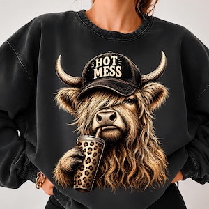May include: A charcoal grey crewneck sweatshirt featuring a graphic of a Highland cow wearing a black baseball cap with the text HOT MESS printed in white. The cow is holding a tumbler with a brown and black leopard print pattern.