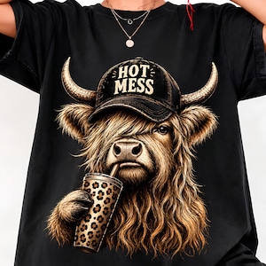 Può includere: Una t-shirt nera con la grafica di una mucca Highland dal pelo lungo che indossa un berretto nero con la scritta "HOT MESS". La mucca tiene in mano un bicchiere con stampa leopardata e una cannuccia. Il disegno è centrato sul petto.