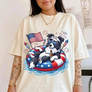 Könnte beinhalten: Cremefarbenes T-Shirt mit einem Cartoon-Hund mit Sonnenbrille und Visier mit der Aufschrift "USA 250", der sich auf einem rot-weiß-blauen Schwimmreifen entspannt. Der Hund hält eine Flasche und ist von Feuerwerk und einer amerikanischen Flagge umgeben.