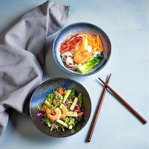May include: Two ceramic bowls with Asian-inspired dishes. One bowl contains noodles, shrimp, vegetables, and mushrooms. The other bowl has a salad with shrimp, greens, and vegetables. Wooden chopsticks are placed on the side.