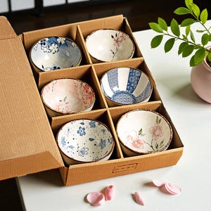 May include: A set of six ceramic bowls with floral designs in a cardboard box. The bowls feature various patterns in blue, pink, and white, with a brown rim. The box is divided into compartments, showcasing the bowls. The overall theme is tableware.