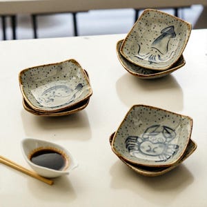 Puede incluir: Una colección de pequeños cuencos cuadrados de cerámica con un esmalte gris moteado y bordes marrones. Cada cuenco presenta una ilustración azul de vida marina, incluyendo un pez, un calamar y un cangrejo. También hay un pequeño cuenco blanco con salsa de soja y palillos.