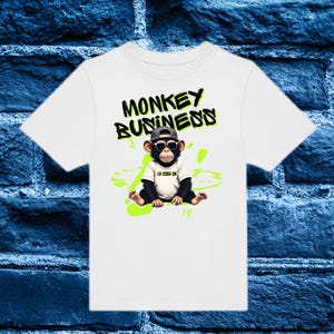 Könnte beinhalten: Weißes T-Shirt mit einem Cartoon-Affen, der eine Sonnenbrille und eine Kappe trägt, mit dem Text "MONKEY BUSINESS" in Schwarz und Grün. Der Affe sitzt auf einem Klecks neongrüner Farbe. Das Shirt hat auch den Text "LV-2203-26".