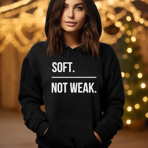 Puede incluir: Sudadera con capucha negra con el texto blanco "SOFT. NOT WEAK.". La sudadera tiene una capucha con cordón y un bolsillo delantero. El fondo está borroso con luces cálidas y doradas.