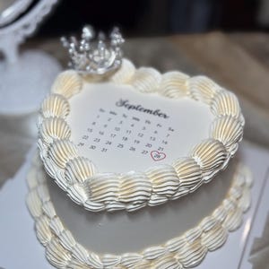 Peut inclure: Un gâteau blanc à deux niveaux avec un glaçage crème et une couronne argentée sur le dessus. La couche supérieure présente un calendrier pour septembre, avec le 28 mis en évidence. Le gâteau est décoré de glaçage en forme de tuyau sur les bords.