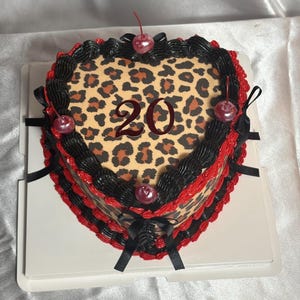 Peut inclure: Gâteau en forme de cœur avec un motif léopard et le chiffre 20 au centre. Le gâteau est décoré de glaçage noir et rouge, de cerises et de rubans noirs. Il repose sur une base en carton blanche.