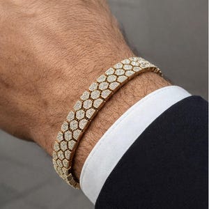 Puede incluir: Una pulsera dorada con diseño de panal, incrustada con pequeñas piedras brillantes. La pulsera se luce en una muñeca, con un traje oscuro y un puño de camisa blanca en el fondo. Una joya de lujo.