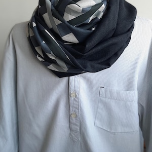 Könnte beinhalten: Ein unendlichkeits-Schal mit geometrischem Muster in Grau-, Weiß- und Marineblautönen. Der Schal ist um den Hals einer Schaufensterpuppe drapiert und zeigt sein Design und seine Textur. Das Hemd ist hellblau gestreift.