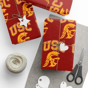 May include: Gift-wrapped presents with a maroon background and a repeating gold USC logo and Trojan head design. The gifts are tied with twine and adorned with white star and heart-shaped tags. A ball of twine, scissors, and blank tags are also present.