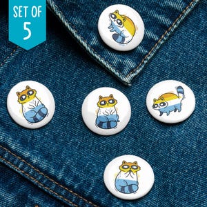 May include: Set of five white round buttons with cartoon raccoons in various poses. Each raccoon has yellow, blue, and brown coloring. The buttons are displayed on a blue denim background. The text "SET OF 5" is in a teal box.