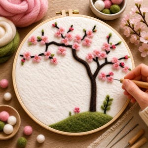 May include: A handmade embroidery hoop with a felted cherry blossom tree design. The tree has pink flowers and a brown trunk, set against a white background with green grass and small pink flowers. The wooden hoop is visible.