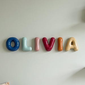 May include: A set of wall-mounted letters spelling the name OLIVIA in a rounded, balloon-like font. Each letter features a distinct glossy colour: dark blue, light blue, pink, dark red, orange, and yellow, arranged horizontally on a light wall.
