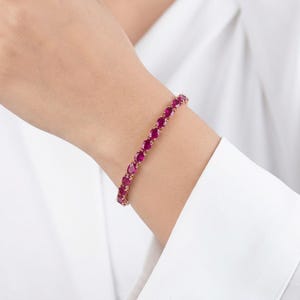 May include: A gold bracelet adorned with oval-cut, deep red gemstones. The bracelet is worn on a wrist, set against a white background. The gemstones are closely set, creating a continuous line of color.