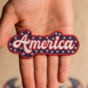 May include: An embroidered patch featuring the word America in white cursive script. The background is dark blue with a pattern of small white stars, all outlined by a thick red border. The patch has a rounded, irregular shape following the text.