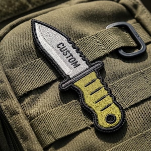 May include: An embroidered patch shaped like a knife with a silver blade featuring the word CUSTOM in black letters and a yellow handle with black stitching details. The patch has a thick black border and is attached to an olive green fabric surface.