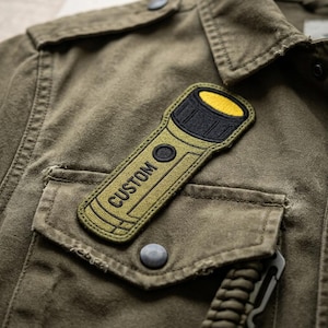 May include: An olive green embroidered patch shaped like a flashlight attached to an olive green jacket. The patch features a yellow lens, a black bezel, a black power button, and the word CUSTOM stitched in black thread along the handle.