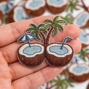 May include: Embroidered patch featuring two coconut halves with blue water, palm trees, and a blue and white umbrella. The patch has brown, green, and blue colors, and a straw is in one coconut. The patch is a tropical theme.