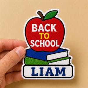 May include: A colorful patch featuring a red apple with the words "BACK TO SCHOOL" in white and yellow. Below the apple are stacked books, and the name "LIAM" is displayed in blue on a white background.