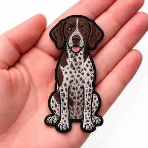 May include: An embroidered iron-on patch featuring a German Shorthaired Pointer dog. The dog has a brown head, brown ears, and a white body covered in brown spots. The dog is sitting with its mouth open, showing a pink tongue and brown eyes.