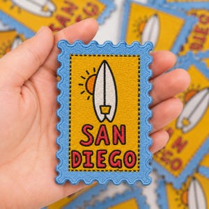 May include: A rectangular embroidered patch shaped like a postage stamp with a light blue scalloped border. The center features a yellow background with a white surfboard, an orange sun, and the red text SAN DIEGO printed in a bold font.