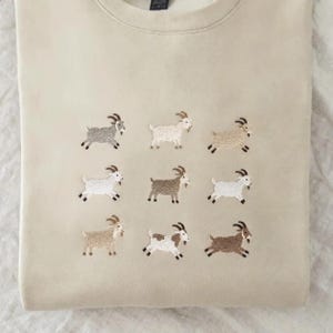 Puede incluir: Una sudadera de cuello redondo en color beige con una cuadrícula bordada de nueve cabras en varios tonos de marrón, blanco y gris. Cada cabra se muestra de perfil, exhibiendo diferentes colores y patrones de pelaje en la prenda.