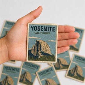 May include: A rectangular embroidered patch featuring a landscape of Half Dome in Yosemite, California. The design includes a blue sky, tan rock formation, and green pine trees. The text YOSEMITE CALIFORNIA is stitched in dark blue at the top.