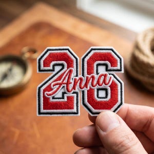 May include: A white patch with the number "26" in red, outlined in black, and the name "Anna" in red script. The patch is held in a hand, with a compass and rope in the background.