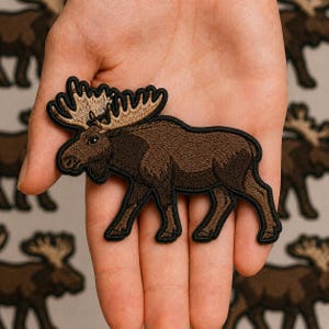 May include: An embroidered iron-on patch shaped like a moose. The moose is dark brown with a tan-colored set of antlers and is outlined in a thick black border. The design features detailed stitching to represent the fur and the shape of the animal.
