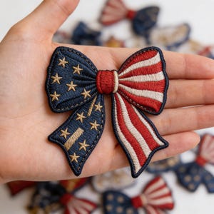 May include: Embroidered bow patch featuring the American flag design. The bow has a navy blue section with gold stars and a red and white striped section. The patch is held in a hand, with other similar patches visible in the background.