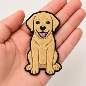 May include: An embroidered patch featuring a tan Labrador retriever puppy sitting in a front-facing position. The dog has floppy ears, a black nose, and an open mouth with a visible pink tongue. The entire design is outlined in a thick black border.