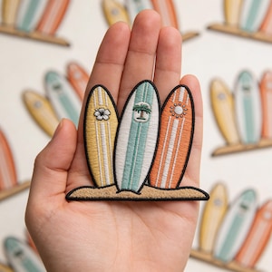 May include: Embroidered patch featuring three surfboards in yellow, teal, and orange, each with unique designs: a flower, a palm tree, and a sun. The surfboards are set on a sandy base, evoking a beach theme. Additional surfboard patches are visible in the background.