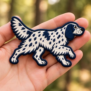 May include: An embroidered patch shaped like a dog in a walking stance. The dog has cream-colored fur with dark navy blue spots and a solid navy blue head and ears. The edges of the patch are finished with a thick navy blue embroidered border.